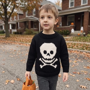 May include: A black long-sleeved knit jumper featuring a large white skull and crossbones graphic on the front, paired with dark grey jeans and an orange fabric pumpkin-shaped treat bag with green leaf accents.