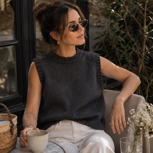 May include: A dark charcoal gray sleeveless knit sweater vest featuring a ribbed crew neckline and ribbed armholes, paired with light cream-colored trousers. The garment has a simple, clean-cut design with a smooth, uniform knit texture.