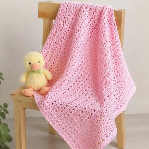 May include: A light pink crocheted baby blanket featuring an openwork diamond pattern with a textured border, accompanied by a small, plush yellow duckling toy with a light green ribbon tied around its neck, both resting on a wooden chair.