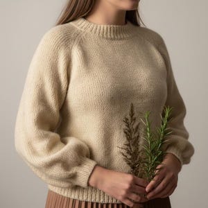 May include: A beige knit sweater with a crew neck and long, slightly puffed sleeves. The sweater is made of a soft, textured yarn. The person is holding a sprig of green and brown plants.