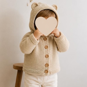 May include: A beige knitted cardigan featuring a hood with two small bear ears on top. The garment has long sleeves with ribbed cuffs, a ribbed hem, and a front button closure with five round wooden buttons. The texture is a classic knit stitch.