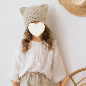 Può includere: Un berretto in maglia beige con due orecchie a punta da gatto sulla parte superiore, abbinato a una camicetta bianca a maniche lunghe con pieghe verticali e abbottonatura frontale, indossato con pantaloni color marrone chiaro con coulisse.