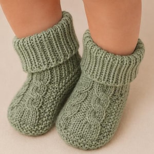 May include: A pair of sage green knitted booties featuring a cabled pattern on the front and a ribbed fold-over cuff at the top. The booties are constructed with a textured knit stitch throughout the foot and heel area, providing a soft appearance.