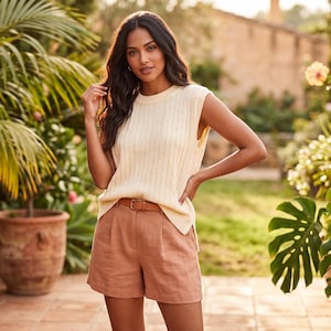 May include: A cream-colored sleeveless knit sweater with a ribbed texture and a crew neckline, paired with solid tan high-waisted shorts featuring a front pleat detail and a thin brown leather belt with a gold-toned buckle at the waist.