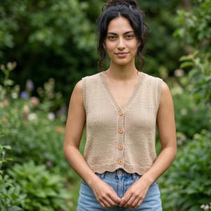 Puede incluir: Un cárdigan beige sin mangas de punto con cuello en V y botones de madera. El cárdigan se lleva sobre unos vaqueros azules. El fondo es un jardín borroso con follaje verde y flores coloridas.