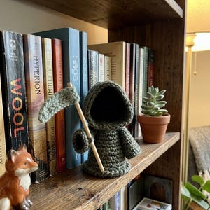 Puede incluir: Una figura de la Muerte hecha a mano con ganchillo verde oliva, sosteniendo una guadaña de madera. La figura se exhibe en una estantería de madera con varios libros y una pequeña suculenta en maceta. También se ve una figura de zorro de cerámica.
