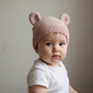 May include: A pale pink knitted baby hat with two small ear-shaped details. The hat is worn by a baby in a white onesie. The background is a plain, neutral colour.