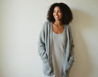 May include: A light gray open-front knit cardigan featuring long sleeves, a ribbed trim along the front opening, and two large patch pockets on the front, worn over a simple light gray sleeveless dress.