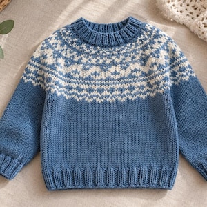 May include: A long-sleeve knit jumper in a medium blue colour featuring a white fair isle pattern around the yoke, neckline, and shoulders. The garment has a ribbed crew neckline, ribbed cuffs, and a ribbed hem, constructed with a stockinette stitch.
