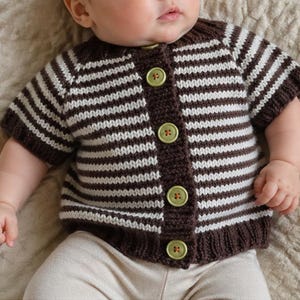 May include: A brown and white striped baby cardigan with short sleeves and green buttons. The knitted sweater is buttoned up and appears to be handmade. The baby is wearing beige trousers.