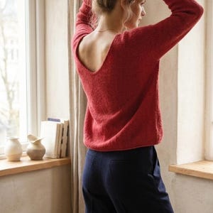 May include: A red sweater with a scoop back is paired with navy blue pants. The sweater has long sleeves and a textured appearance. The model is standing near a window, with natural light illuminating the scene. The outfit is simple and elegant.