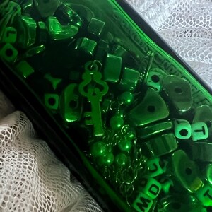 May include: A green glass bottle filled with various green objects, including beads, a key charm, and lettered dice. The bottle's contents are illuminated, creating a vibrant display. The background features a white, lacy fabric.