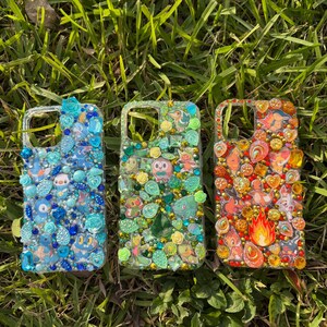 May include: Three rectangular phone cases decorated with various small character stickers and assorted blue, green, and orange rhinestones, gems, and beads. The left case features blue tones, the center case features green tones, and the right case features orange and red tones.