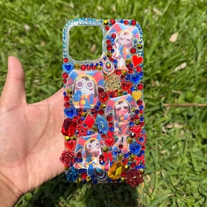 Puede incluir: Funda de teléfono decorada con un diseño de personaje de dibujos animados, adornada con pedrería roja, azul y amarilla, corazones y rosas. La funda tiene un borde transparente con pedrería azul.