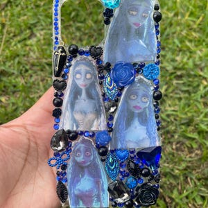 Puede incluir: Funda de teléfono decorativa con imágenes de un personaje de piel azul con pelo largo y blanco. La funda está adornada con pedrería, cuentas y abalorios azules y negros, incluyendo un ataúd y rosas. La funda tiene un fondo transparente.