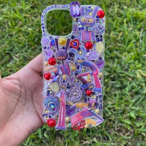 Puede incluir: Funda de teléfono transparente decorada con pedrería morada, roja y dorada, y personajes de dibujos animados. La funda presenta una variedad de adornos, incluyendo corazones, flores y gemas. Los personajes de dibujos animados están en varias poses y atuendos.