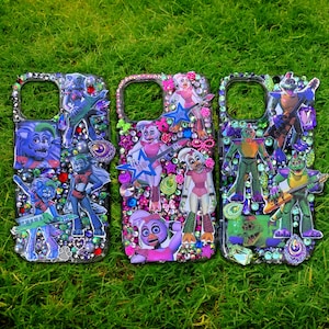 May include: Three bedazzled phone cases with character designs. The cases feature colorful rhinestones and various character cutouts. The cases are on a green grassy surface. The cases have a clear background.