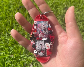 May include: A red motel-style keychain with a silver key ring. The keychain is decorated with playing card suits, dice, and a picture of a woman in a costume. The keychain is embellished with red and black beads.
