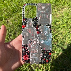 Puede incluir: Una funda de teléfono con imágenes de figuras militares con equipo de combate. La funda está adornada con cuentas negras y rojas y pequeños detalles de rosas rojas. La funda se sostiene sobre un fondo de hierba verde.