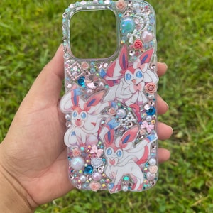 May include: Clear phone case decorated with Sylveon characters, pink and blue gems, pearls, and small pink roses. The case has a clear border with embedded rhinestones. The design is set against a background of green grass.
