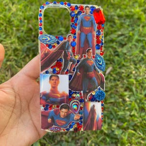 May include: Clear phone case decorated with images of Superman in various poses. The case is embellished with red, blue, and clear rhinestones around the edges and between the images. The background is green grass.