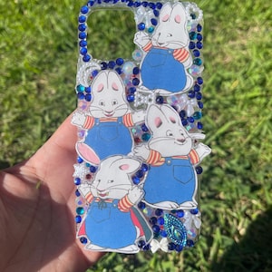 May include: A clear phone case decorated with blue rhinestones and cartoon images of a white rabbit wearing blue overalls. The case is embellished with white pearls and other colorful gems.