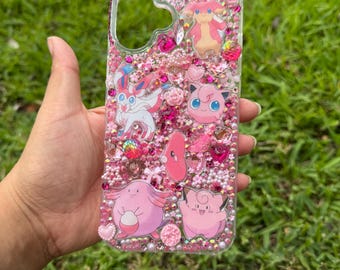 Puede incluir: Una funda de teléfono transparente decorada con cuentas, cristales y pequeños dijes de color rosa y blanco. La superficie presenta varias pegatinas de personajes de dibujos animados rosas, incluyendo a Sylveon, Jigglypuff, Chansey y Clefairy.