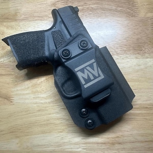 Puede incluir: Funda de pistola negra con un clip y las letras "MV" grabadas en el lateral.