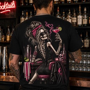 May include: A black t-shirt featuring a graphic print of a skeleton with a messy bun, pink headband, and sunglasses, sitting at a bar. The skeleton holds a pink cocktail and a cigarette, with a skull, shaker, and lime wedge on the bar counter.