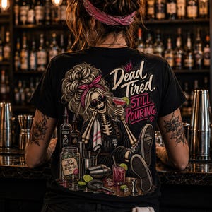 May include: A black t-shirt featuring a graphic of a skeleton bartender with a pink headband and sunglasses, holding a cocktail. The design includes the text 'Dead Tired. STILL POURING', a tip jar labelled 'TIPS KEEP ME ALIVE-ISH', and various bar tools.