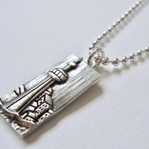 May include: Silver necklace with a rectangular pendant featuring a detailed depiction of a city skyline.