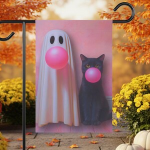 May include: A Halloween garden flag featuring a ghost and a black cat, both with pink bubble gum bubbles. The ghost is draped in a white sheet with black eye holes. The cat has yellow eyes. The background is pink.