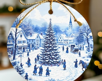 May include: Round Christmas ornament featuring a winter village scene in shades of blue and white. A large decorated Christmas tree is the focal point, surrounded by people and buildings. The ornament is suspended by a gold-colored string.