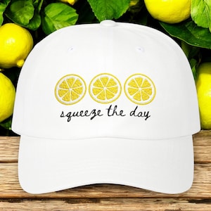 May include: White baseball cap featuring three embroidered yellow lemon slices above the phrase "squeeze the day" in black script. The cap is set against a background of lemons and green leaves.