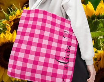 May include: A pink and white checkered tote bag with the name "Olivia" in a cursive font. The bag is held by a person wearing a white hoodie. The background features sunflowers.