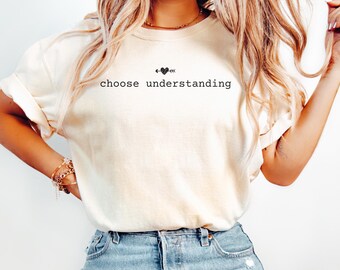 May include: Cream-colored t-shirt with the phrase "choose understanding" in a black, simple font. Above the text is a small graphic of a heart with an arrow. The shirt is paired with blue jeans.