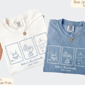May include: Two t-shirts, one ivory and one blue jean, with a matching graphic print. The print features a bikini, cocktail, and beach umbrella design, with the text "Helen's Bachelorette, Miami, Florida". Each shirt has a gold necklace.
