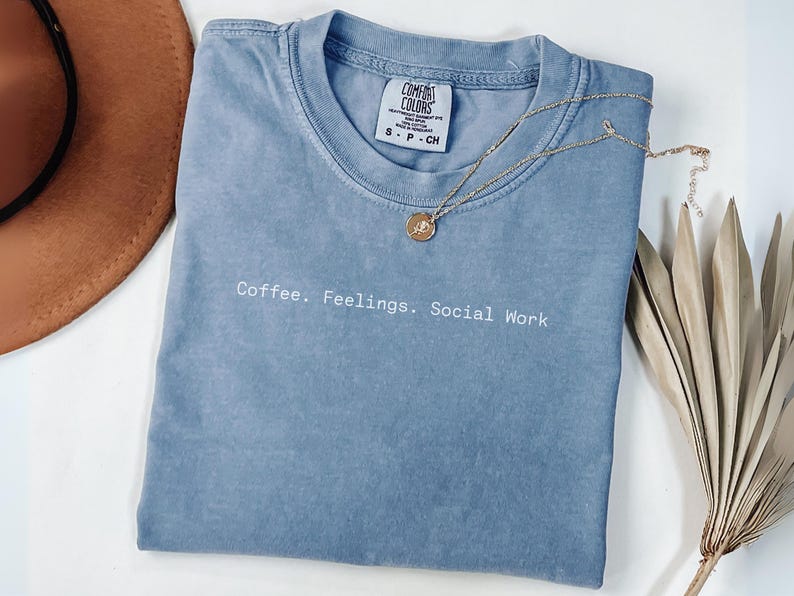 May include: A blue Comfort Colors t-shirt with the text "Coffee. Feelings. Social Work" in white. A gold necklace with a pendant rests on the shirt. A brown hat and dried plant are also in the image.