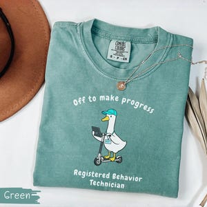 May include: Light green Comfort Colors t-shirt with the text "Off to make progress" and a cartoon duck riding a scooter. The shirt also says "Registered Behavior Technician". A brown hat and a necklace are also in the image.