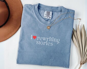 May include: A blue Comfort Colors t-shirt with the phrase "I ❤️ rewriting stories" in white script. A gold necklace with a pendant rests on the shirt. A brown hat and dried plant are in the background.