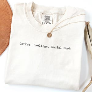 May include: Cream-colored t-shirt with the text "Coffee. Feelings. Social Work" in black. A gold necklace with a circular pendant rests on the shirt. A brown hat and dried plant are in the background.