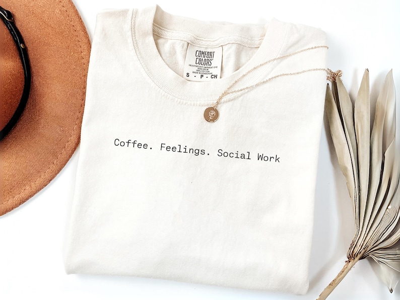 May include: Cream-colored t-shirt with the text "Coffee. Feelings. Social Work" in black. A gold necklace with a circular pendant rests on the shirt. A brown hat and dried plant are in the background.