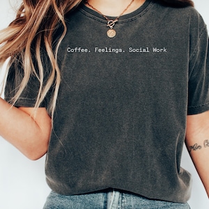 May include: Dark gray t-shirt with the text "Coffee. Feelings. Social Work" in a white, sans-serif font. The shirt has a slightly faded appearance, suggesting a vintage or worn look. The overall style is casual and comfortable.