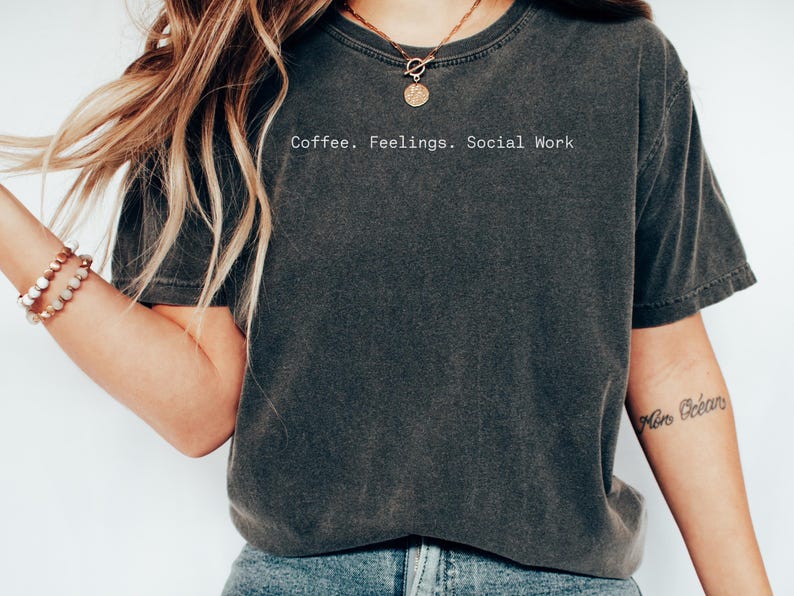 May include: Dark gray t-shirt with the text "Coffee. Feelings. Social Work" in a white, sans-serif font. The shirt has a slightly faded appearance, suggesting a vintage or worn look. The overall style is casual and comfortable.
