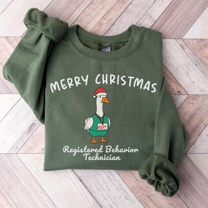 May include: Green sweatshirt with the words "MERRY CHRISTMAS" above a cartoon goose wearing a Santa hat and a name tag that says "RBT". Below the goose is the text "Registered Behavior Technician".