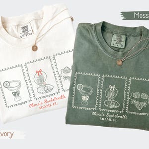 May include: Two t-shirts, one ivory and one moss green, are displayed. Each shirt features a graphic of cocktail glasses, seashells, and lingerie, with the text "Olivia's Bachelorette, Miami, FL." Gold necklaces with round pendants are draped on the shirts.