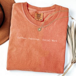 May include: A coral-colored t-shirt with the text "Coffee. Feelings. Social Work" in white. A gold necklace with a pendant and a brown hat are also visible. The shirt is made of a soft material.
