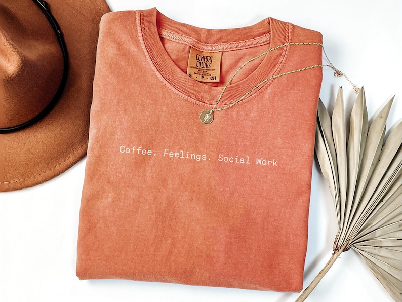 May include: A coral-colored t-shirt with the text "Coffee. Feelings. Social Work" in white. A gold necklace with a pendant and a brown hat are also visible. The shirt is made of a soft material.