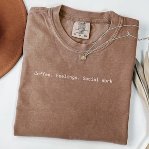 May include: A brown Comfort Colors t-shirt with the text "Coffee. Feelings. Social Work" in white. A silver necklace with a pendant and a brown hat are also visible. A dried plant is on the right.
