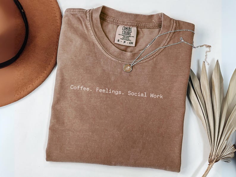 May include: A brown Comfort Colors t-shirt with the text "Coffee. Feelings. Social Work" in white. A silver necklace with a pendant and a brown hat are also visible. A dried plant is on the right.